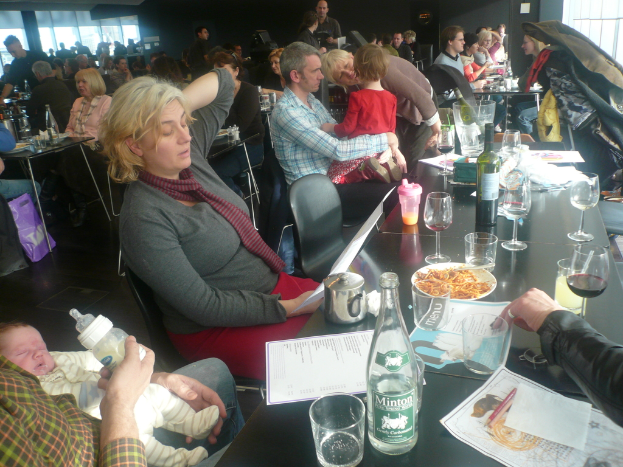 Ein Innenblick in ein belebtes Restaurant mit Menschen an Tischen, die Gläser, Teller mit Essen, Getränkeflaschen, Speisekarten und andere Gegenstände haben.