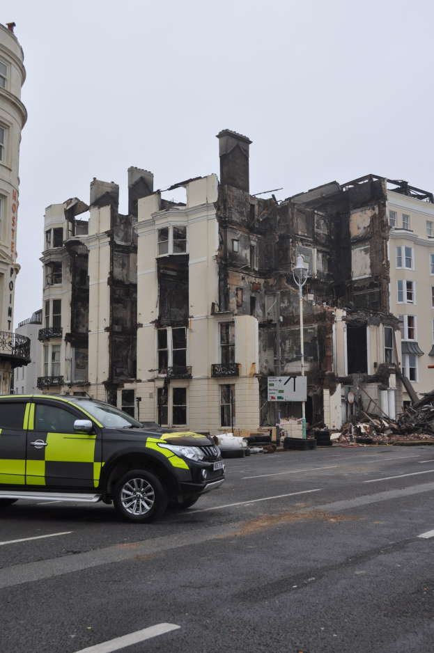 Polizeiauto vor einem schwer beschädigten Gebäude mit zersplitterten Fenstern und verstreuter Trümmer geparkt, andere Gebäude und Himmel im Hintergrund sichtbar.