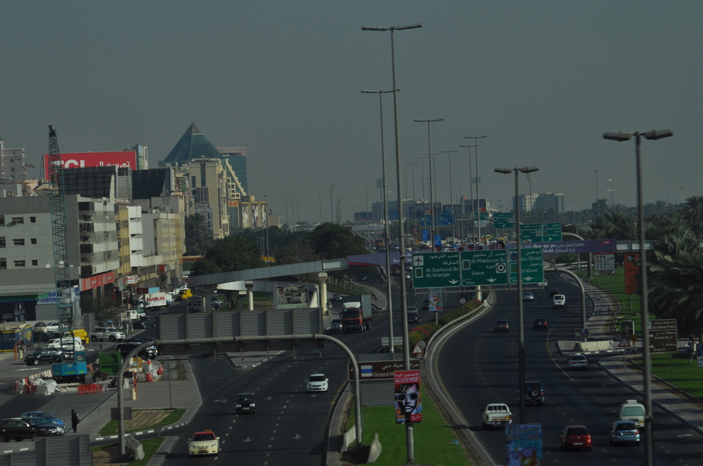 Eine Luftaufnahme einer Stadt mit Gebäuden, Türmen, Fahrzeugen, Werbetafeln, Schildern, Straßen, Bäumen, Straßenlaternen, Menschen, Verkehrskegeln und Informationsanzeigen unter einem klaren Himmel.