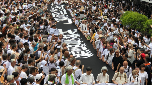 Eine große Gruppe von Menschen marschiert auf der Straße, hält Protestschilder und Banner in der Hand, mit Grünzeug auf der rechten Seite und einem Gebäude im Hintergrund.