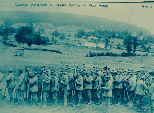Eine Gruppe deutscher Gefangener in Frankreich mit Waffen im Freien stehend, umgeben von Bäumen, Häusern und einem klaren blauen Himmel.