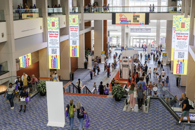 Eine große Gruppe von Menschen geht durch ein großes Gebäude mit Bannern, Topfpflanzen, Rolltreppen, Geländern, Säulen, Texttafeln, Glastüren und anderen Gegenständen, einige tragen Taschen, wahrscheinlich beim Nationalen Buchfestival.