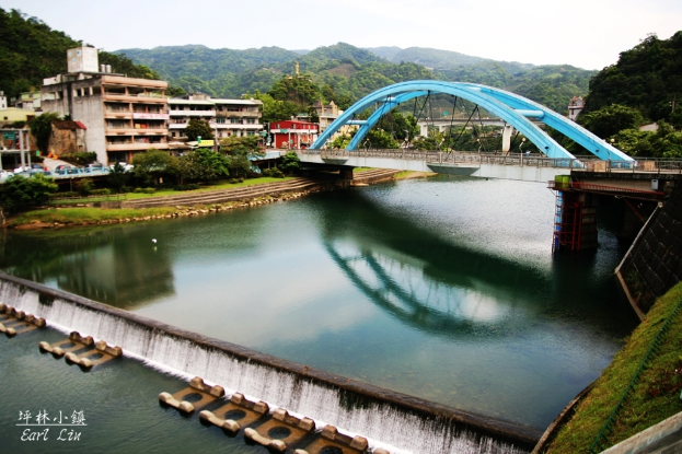 Eine Brücke überspannt einen Fluss mit Gebäuden, Bäumen, Boden, Hügeln, Zäunen und Himmel im Hintergrund.