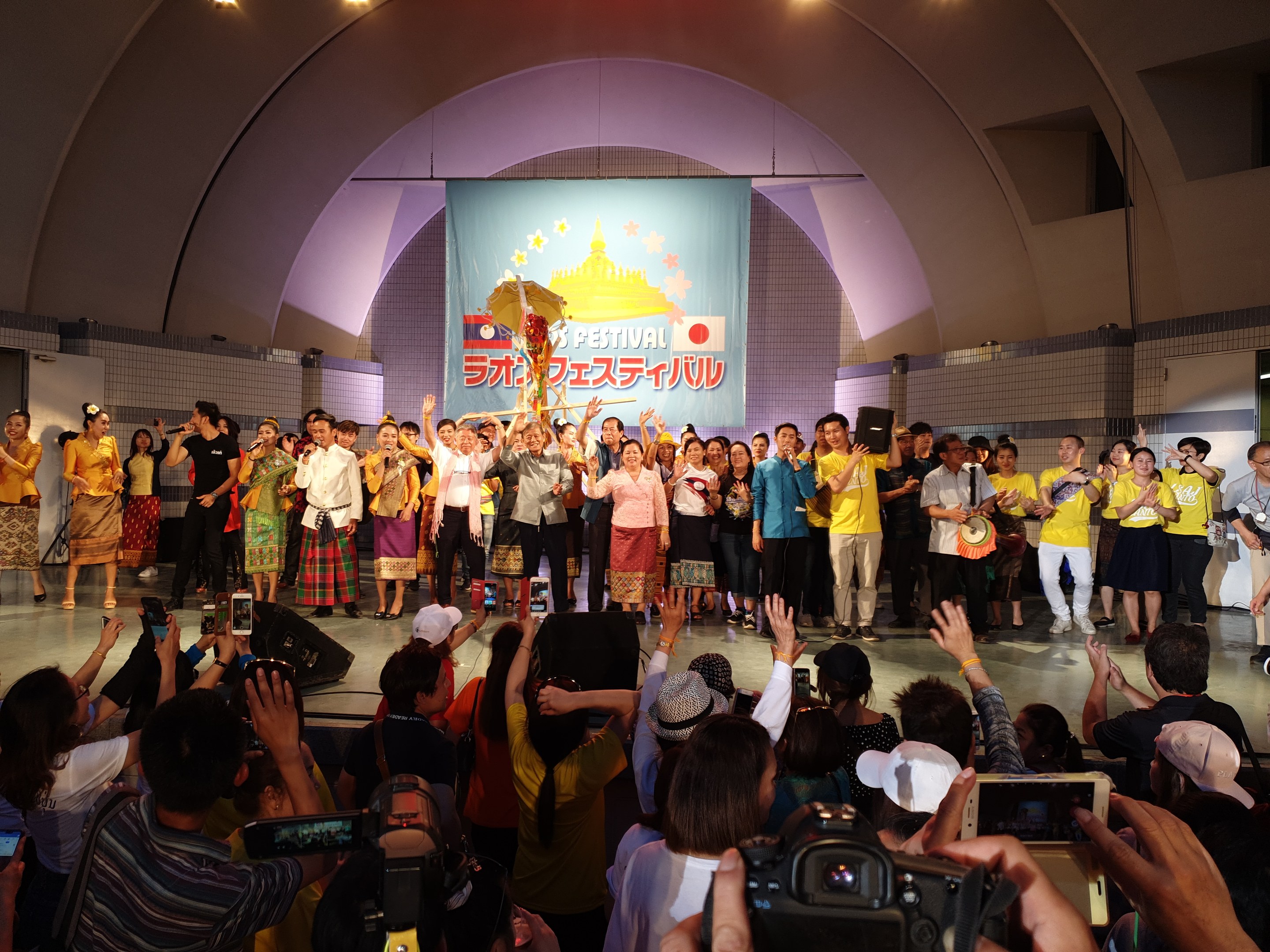 Eine Gruppe von Menschen auf einer Bühne mit Kameras, Handys und Musikinstrumenten während einer Festivaleröffnung mit Bannern, Wänden und Deckenlampen im Hintergrund.