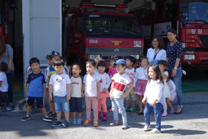 Gruppe von Kindern vor einem Feuerwehrauto an einer Feuerwache, einige tragen Mützen, mit weiteren Feuerwehrautos im Hintergrund.