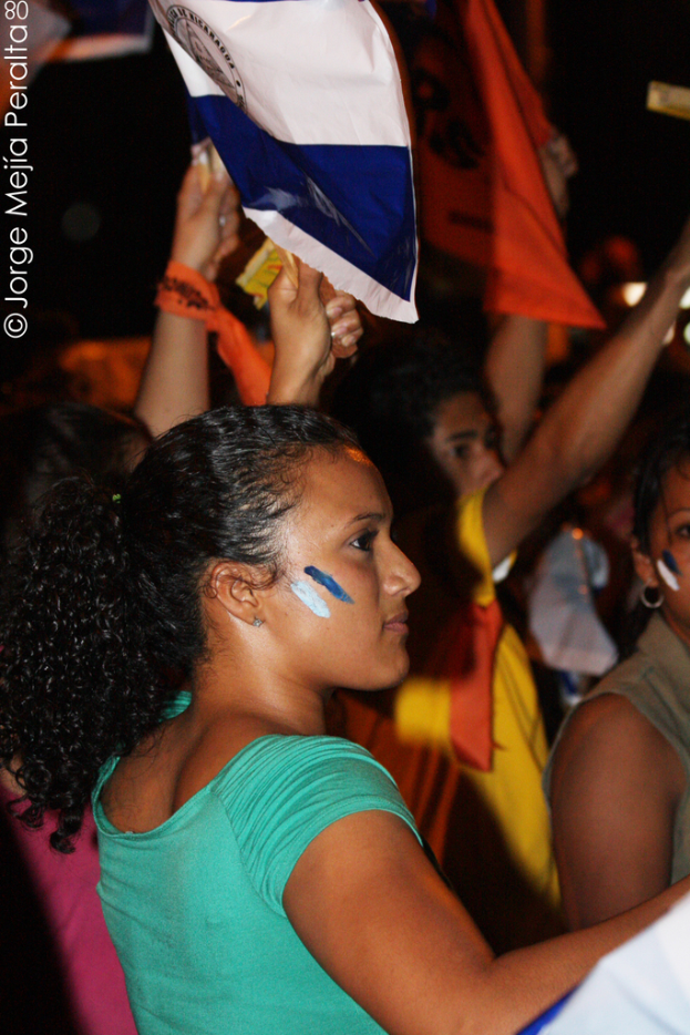 Eine Frau in einem gr√ľnen T-Shirt steht auf einer Demonstration und h√§lt ein weißes Schild in der Hand, hinter ihr stehen viele andere Menschen.