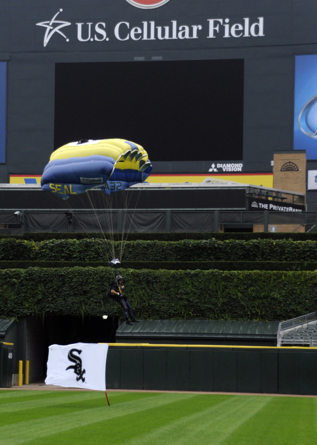 Eine Person paragleitet über ein Baseballfeld mit einem Fallschirm, umgeben von Grünflächen, einem Banner, Tafeln mit Text und Logos sowie einem Gebäude im Hintergrund.