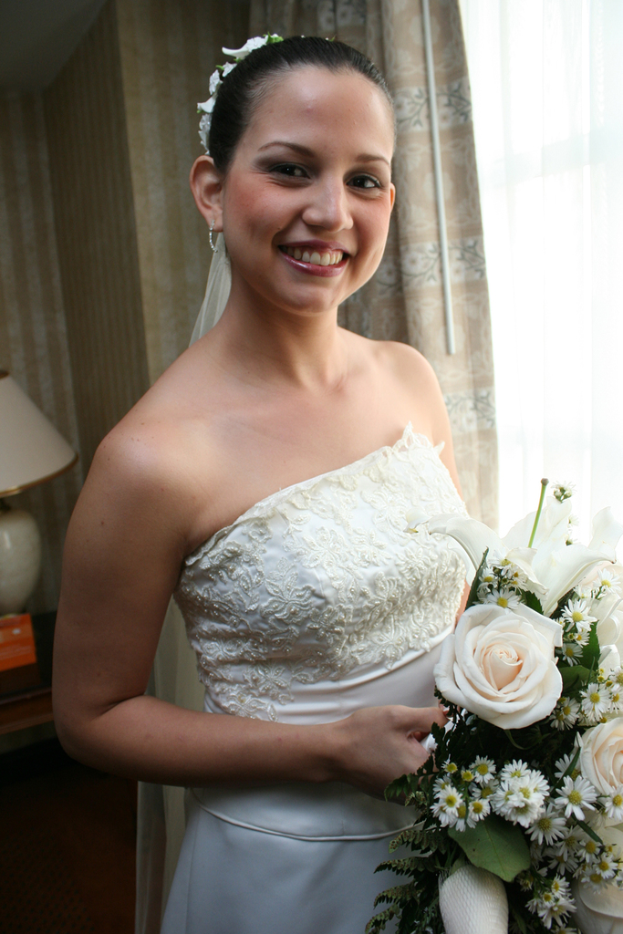 Eine Braut steht strahlend in der Mitte, hält einen Blumenstrauß und hat im Hintergrund einen Vorhang und eine Wand sowie eine Lampe auf einem Tisch links.