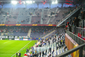 Ein großes Stadion voller Zuschauer, die ein Fußballspiel sehen, mit einigen Menschen, die auf Stühlen auf dem Feld stehen und sitzen, einem Geländer und einer Fahne auf der rechten Seite und einem leeren Stadion mit Deckenlampen im Hintergrund.