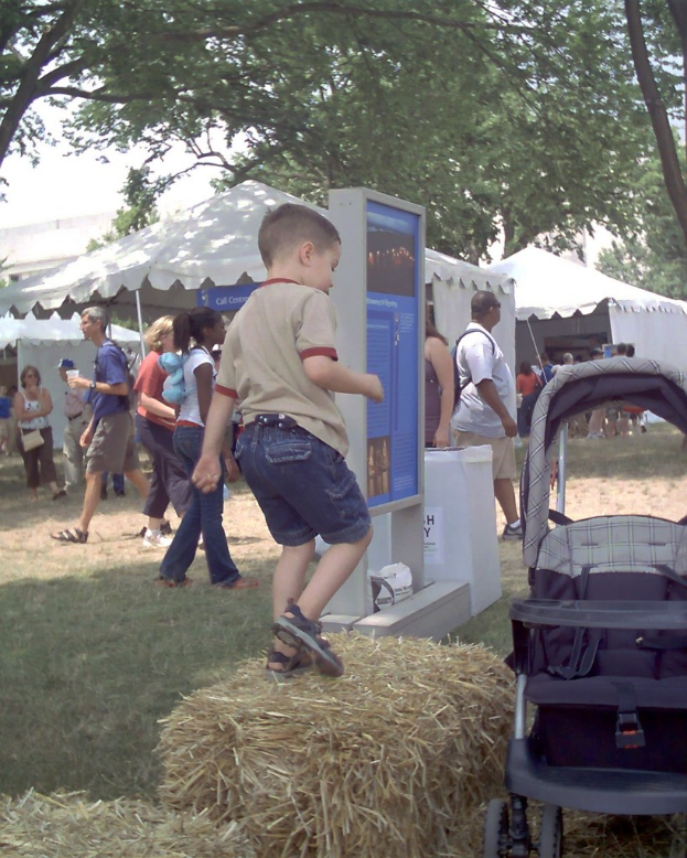 Eine Szene mit mehreren Zelten und Menschen, die darum herumgehen, ein Anzeigebrett vorn, trockenes Gras mit einem spielenden Kind und Bäume auf beiden Seiten.