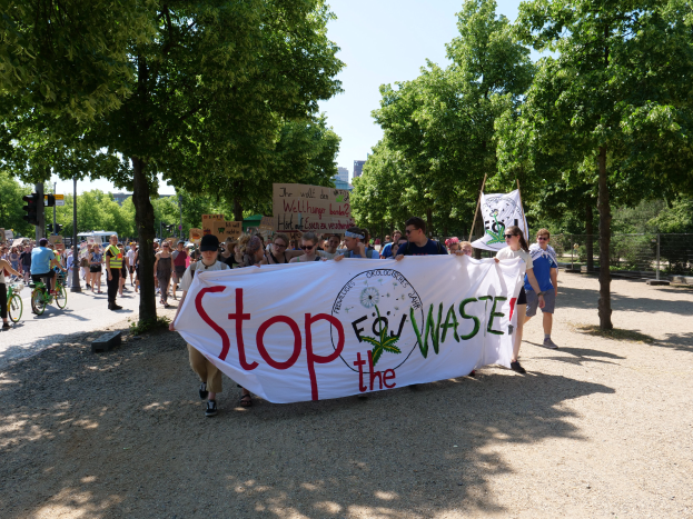 Eine Gruppe von Menschen marschiert eine stra├čen entlang, die ein "Stop the Waste"-Plakat und verschiedene Plakate h├Ąlt, einige fahren Fahrr├Ąder, Geb├Ąude im Hintergrund unter einem klaren blauen Himmel.
