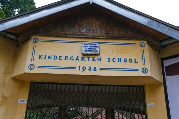Ein gelbes Gebäude mit einer "Kindergarten-Schule"-Schilder und einer Namenskarte, mit einem Gitter unten und Bäumen vor einem klaren blauen Himmel im Hintergrund.