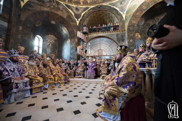 Eine Gruppe von Menschen sitzt in einer Kirche, wobei eine Person rechts mit einer Kamera steht, andere auf einer Treppe, Geländern, Fenstern im Hintergrund, Gemälde an den Wänden, eine Deckenlampe von der Decke hängend und Text mit einem Logo in der rechten unteren Ecke, alle tragen Kronen.