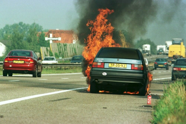 Ein Auto in Flammen auf dem Seitenstreifen, umgeben von anderen Fahrzeugen, mit Bäumen, Gebäuden und einem klaren blauen Himmel im Hintergrund; Gras und ein Feuerlöscher sind auf der rechten Seite zu sehen.
