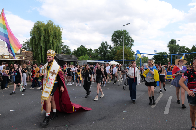 Eine Gruppe von Menschen marschiert auf der 2018er Gay Pride Parade, einige mit Musikinstrumenten und Mützen, eine Regenbogenfahne tragend, während sie eine Straße mit Laternenpfählen, Bäumen, Hütten und einem bewölkten Himmel im Hintergrund entlanggehen.