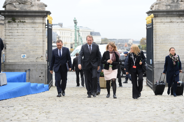 Eine Gruppe von Mitarbeitern geht in einen Bereich mit einem Tor, hinter dem sich viele Fahrzeuge und Gebäude befinden, während eine Frau rechts zwei Koffer trägt.