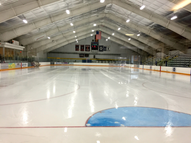 Große Indoor-Eisfläche mit Deckenbeleuchtung, Texttafeln, einer Wand, Treppen mit Geländern und einigen Menschen, die einem Hockey-Sportplatz ähneln.