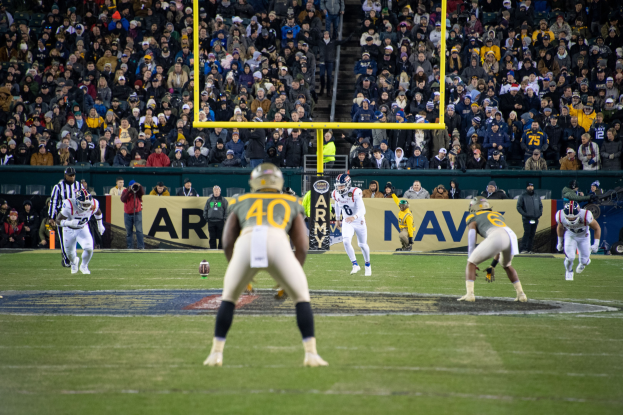 Ein Fußballspiel wird in einem Stadion gespielt, mit Spielern in Sportbekleidung und Helmen im Vordergrund und einer Zuschauermenge im Hintergrund sowie sichtbaren Pfosten und Tafeln mit Text.
