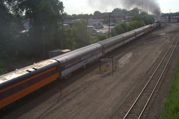 Ein Zug auf Schienen mit Bäumen, Strommasten mit Kabeln, Autos und Gebäuden links, eine Brücke vorne und aus dem Zug austretender Rauch.