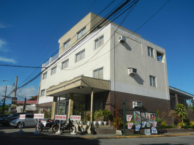 Wohnanlage mit parkenden Autos, Schildern, Strommasten, Bäumen und einem bewölkten Himmel.