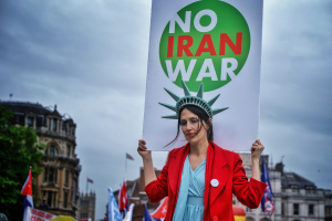 Eine Frau steht in der Mitte und hält ein Schild mit der Aufschrift "Kein Iran-Krieg" mit einem Statue of Liberty-Kopfschmuck, mit Menschen, Flaggen, Gebäuden, Bäumen und einem klaren blauen Himmel im Hintergrund.