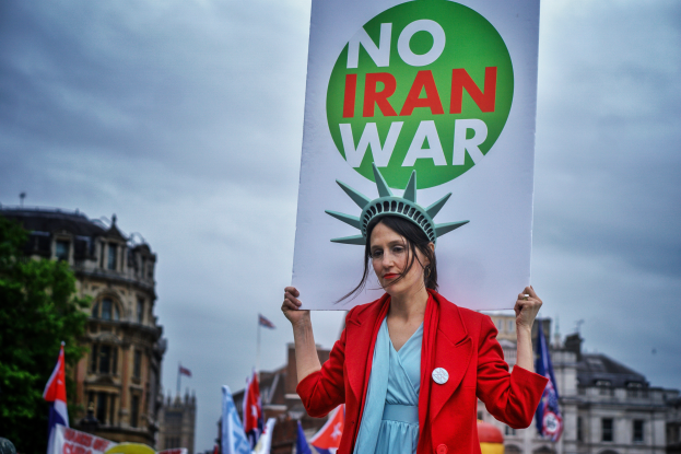 Eine Frau steht in der Mitte und hält ein Schild mit der Aufschrift "Kein Iran-Krieg" mit einem Statue of Liberty-Kopfschmuck, mit Menschen, Flaggen, Gebäuden, Bäumen und einem klaren blauen Himmel im Hintergrund.