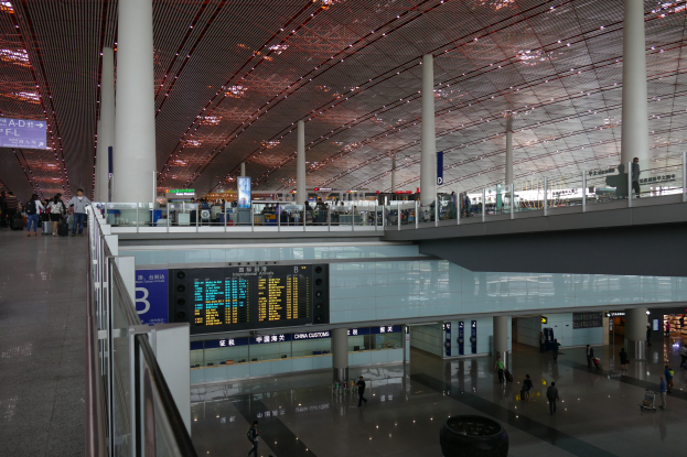 Ein belebter Flughafeninnenraum mit Menschen, die gehen, einige tragen Taschen, umgeben von Geländern, Säulen, Texttafeln und Deckenleuchten.