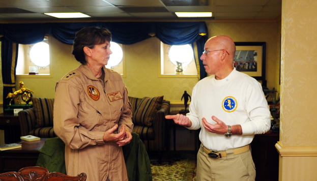 Ein Mann und eine Frau unterhalten sich in einem Wohnzimmer mit Möbeln, Bücherregalen und einem Foto auf dem Wand, mit Fenstern und Lampen, die eine gemütliche Atmosphäre schaffen.