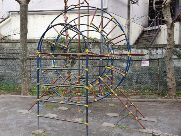 Spielplatz mit großem Metallklettergerüst, umgeben von Bäumen und einer Maschendrahtzaun, mit Treppen mit Geländern, die zu einem Gebäude mit Fenstern und Rohren führen, einem Wegweiser und grasbewachsenem Boden.