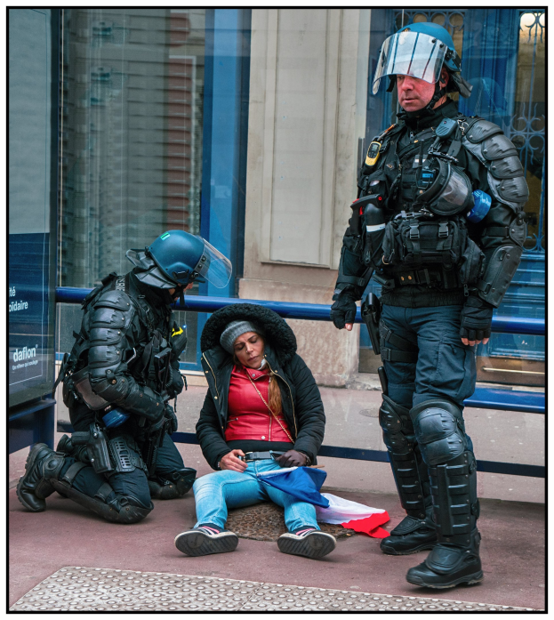 Frau sitzt auf dem Boden neben zwei uniformierten Polizeibeamten, mit einem Gebäude mit Glastüren und einem Schild im Hintergrund.