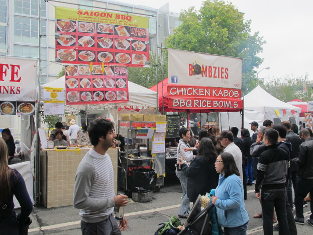 Menschen stehen auf einer Straße vor der Stadt, mit einem Mann in einem grauen und weißen T-Shirt, der eine Wasserflasche hält, und einer Frau in einer blauen Jacke, die einen Kinderwagen schiebt. Hinter ihnen sehen wir Stände mit weißen und roten Zelten, Banner in rot, weiß und gelb mit Text, sowie Gebäude, Bäume und eine Straßenlaterne im Hintergrund.