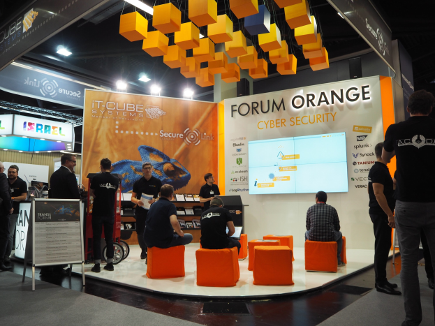 Eine Gruppe von Menschen an einem Messestand für IT-Cube Systems Forum Orange Cyber Security, mit sitzenden und stehenden Teilnehmern, einer Tafel mit Text, einem Bildschirm, Regalen mit Gegenständen, Schildern, Deckenbeleuchtung und Lampen.