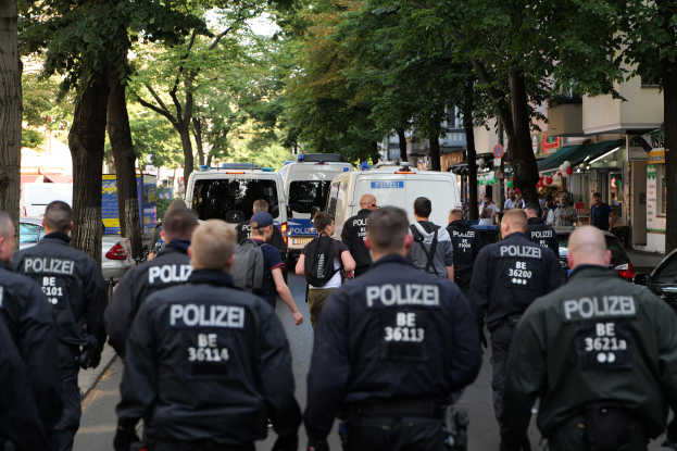 Polizisten gehen eine von Bäumen gesäumte Straße mit geparkten Fahrzeugen und Passanten entlang.