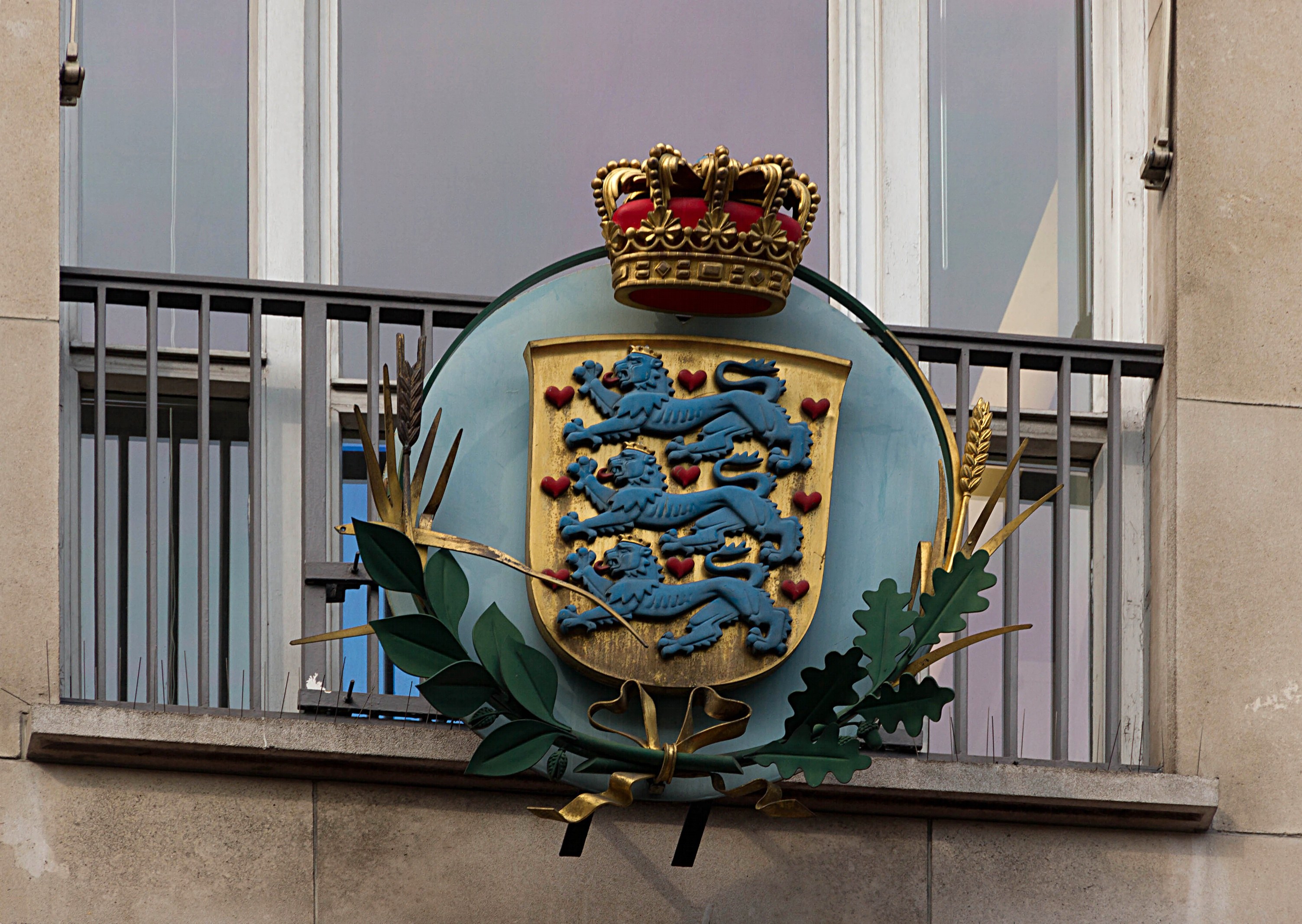 Wappen der britischen Königsfamilie auf einem Gebäude mit einer Krone und einem Blatt-Logo, Fenstern und Gittern im Hintergrund.