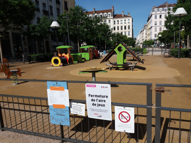 Ein umzäunter Spielplatz auf einem Stadtplatz mit Schildern am Zaun, umgeben von Bäumen, Laternenmasten und Gebäuden unter einem sichtbaren Himmel.