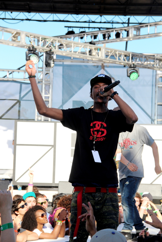 Eine Person in einem schwarzen T-Shirt hält ein Mikrofon auf einer Bühne mit Menschen drumherum und einer weiteren Person in einem grauen T-Shirt im Hintergrund unter einem blauen Himmel.
