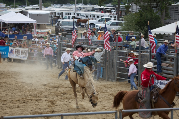 Zwei Menschen reiten auf Pferden bei einem Wettbewerb namens Charreada, umgeben von Gruppen von Menschen, Bannern und Fahnen, Zeltplanen, geparkten Fahrzeugen und Bäumen im Hintergrund.