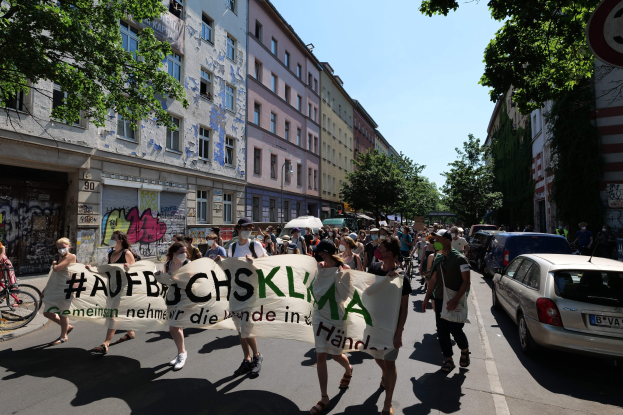 Eine Gruppe von Menschen marschiert mit einem Banner die Straße entlang, mit parkenden Autos auf der rechten Seite, einem Radfahrer auf der linken Seite und Gebäuden, Bäumen und einem Schild im Hintergrund unter einem sichtbaren Himmel.
