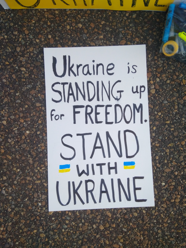 Ein weißes Blatt Papier mit den Worten "Ukraine steht für Freiheit auf, steht mit Ukraine" liegt vor einer gelben Fahne auf dem Boden. Rechts daneben eine Plastikabdeckung mit verschiedenen Gegenständen.