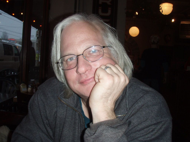 Ein Mann steht mit der Hand auf seinem Gesicht, ein Tisch mit Gläsern und Flaschen links daneben, ein Glas zeigt einen Van auf der Straße und Lichter rechts.