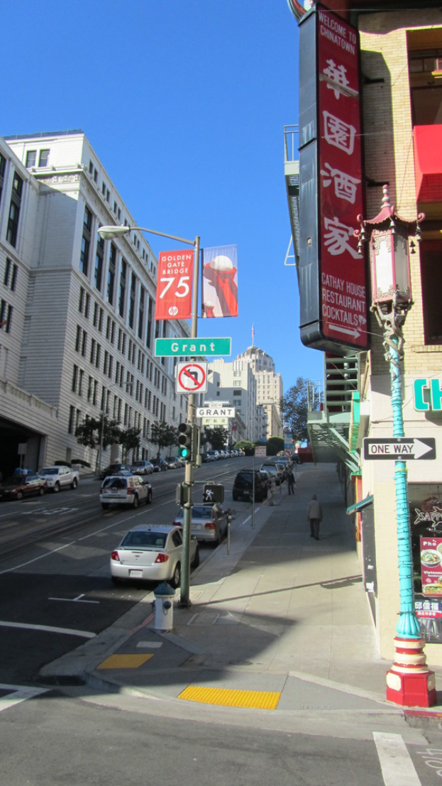 Eine Stadtansicht mit Fahrzeugen auf der Straße, Pfählen und Bäumen auf dem Gehweg, Gebäuden und einem Himmel darüber.