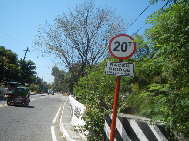 Eine Straße mit fahrenden Fahrzeugen, ein Geschwindigkeitsbegrenzungsschild am Straßenrand, eine Trennlinie, Bäume, Strommasten mit Drähten und einen klaren blauen Himmel im Hintergrund.
