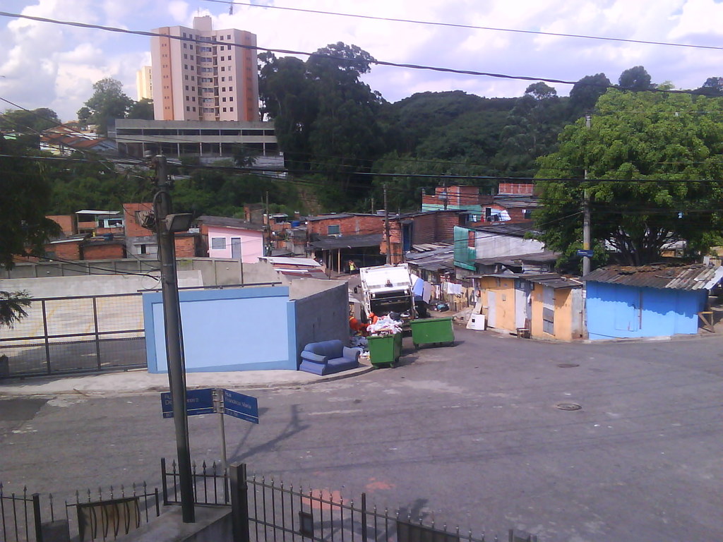 Eine Straßenszene mit Gebäuden, Häusern, Bäumen, Pfählen, Kabeln, einem Schild, Zäunen, Mülltonnen, einem Fahrzeug und Wolken am Himmel.