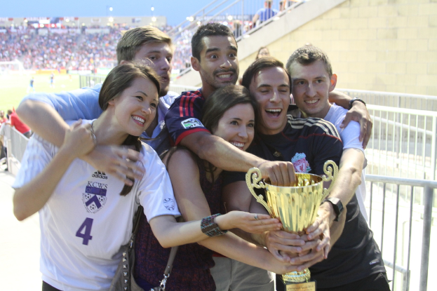 Sechs Menschen, zwei Frauen und vier Männer, die eine Trophäe halten und lächeln, posieren vor einem Eisengeländer für ein Foto, mit einer Wand und einem Geländer oben rechts, Menschen und Lichter im Hintergrund, möglicherweise in einem Stadion.