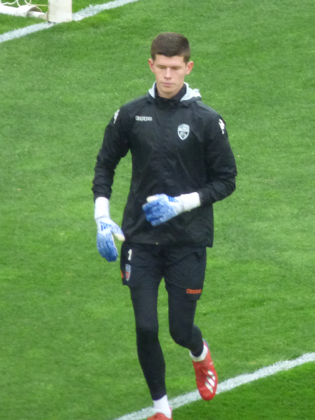 Ein Mann in einer schwarzen Jacke und blauen Handschuhen geht auf einem Fuballfeld mit einem Tor im Hintergrund, wobei er Fuballhandschuhe trägt.