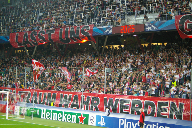 Fußballspiel in einem Stadion mit großer Zuschauermenge, saftig grünem Rasen, Torpfosten links, Bodenbanner, Menschen mit Fahnen, Hintergrundgeländer, Schilder, Lampen und andere Gegenstände.