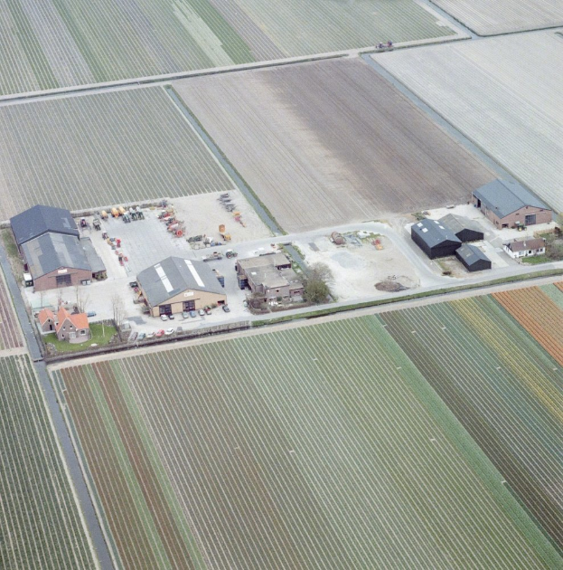 Eine Luftaufnahme eines Bauernhofs in einem Feld, mit Häusern, Fahrzeugen, Bäumen und umliegenden Feldern.