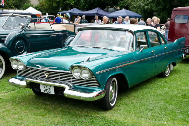 Vintage cars parked on grass with people standing behind them, tents in the background, trees at the top, and a flag in the top left.
