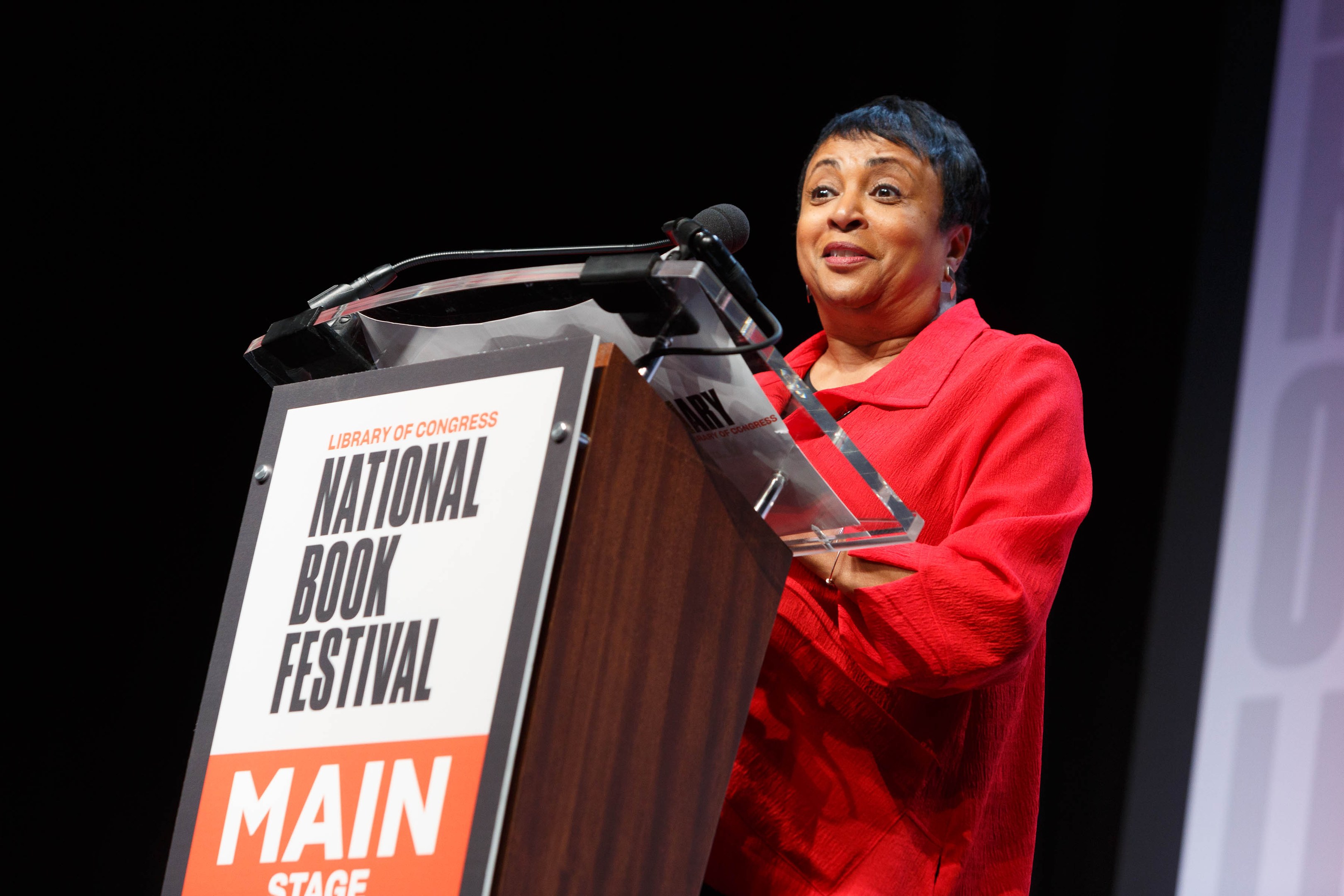 Frau in rotem Kleid, das an einem Pult mit der Aufschrift "National Book Festival" spricht und ein Bildschirm rechts daneben.