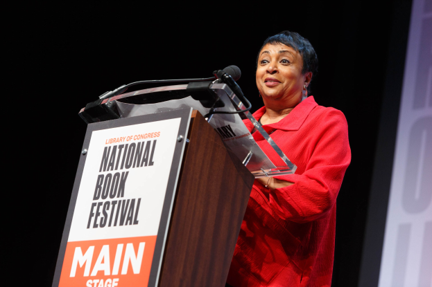 Frau in rotem Kleid, das an einem Pult mit der Aufschrift "National Book Festival" spricht und ein Bildschirm rechts daneben.